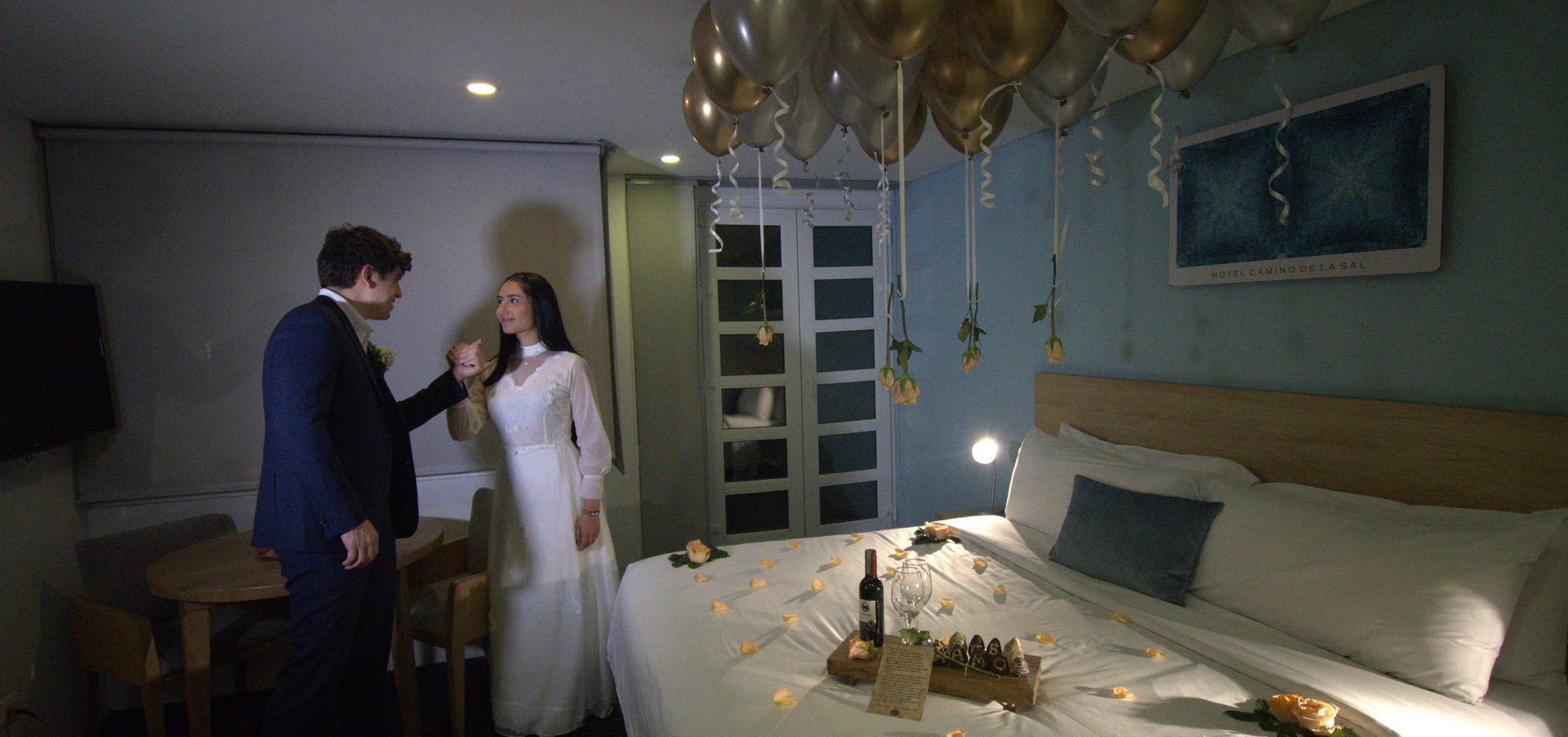 Pareja de recién casados en habitación decorada con globos dorados y plateados con flores colgantes y pétalos de rosa sobre la cama, Hotel Camino de la Sal en Zipaquirá